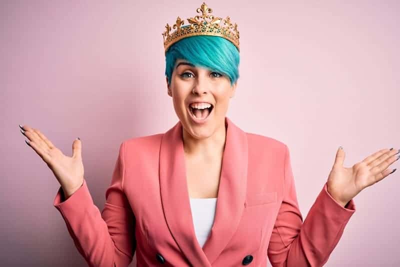 A blue colored hair woman, wearing a pink suit in front of a dark pink background.